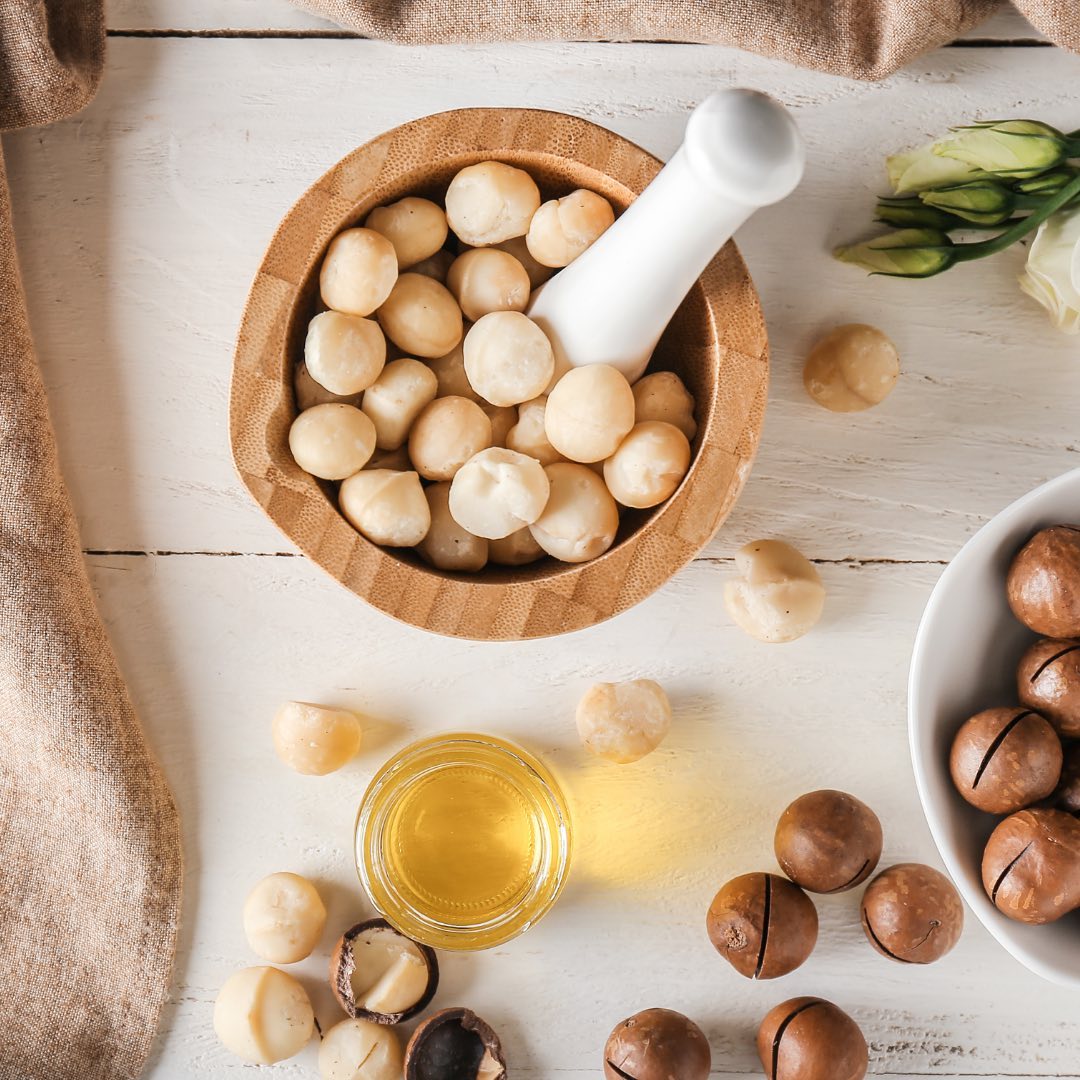 A bowl of shelled macadamia nuts surrounded by whole macadamia nuts in their shells and a bottle of macadamia oil on a wooden table