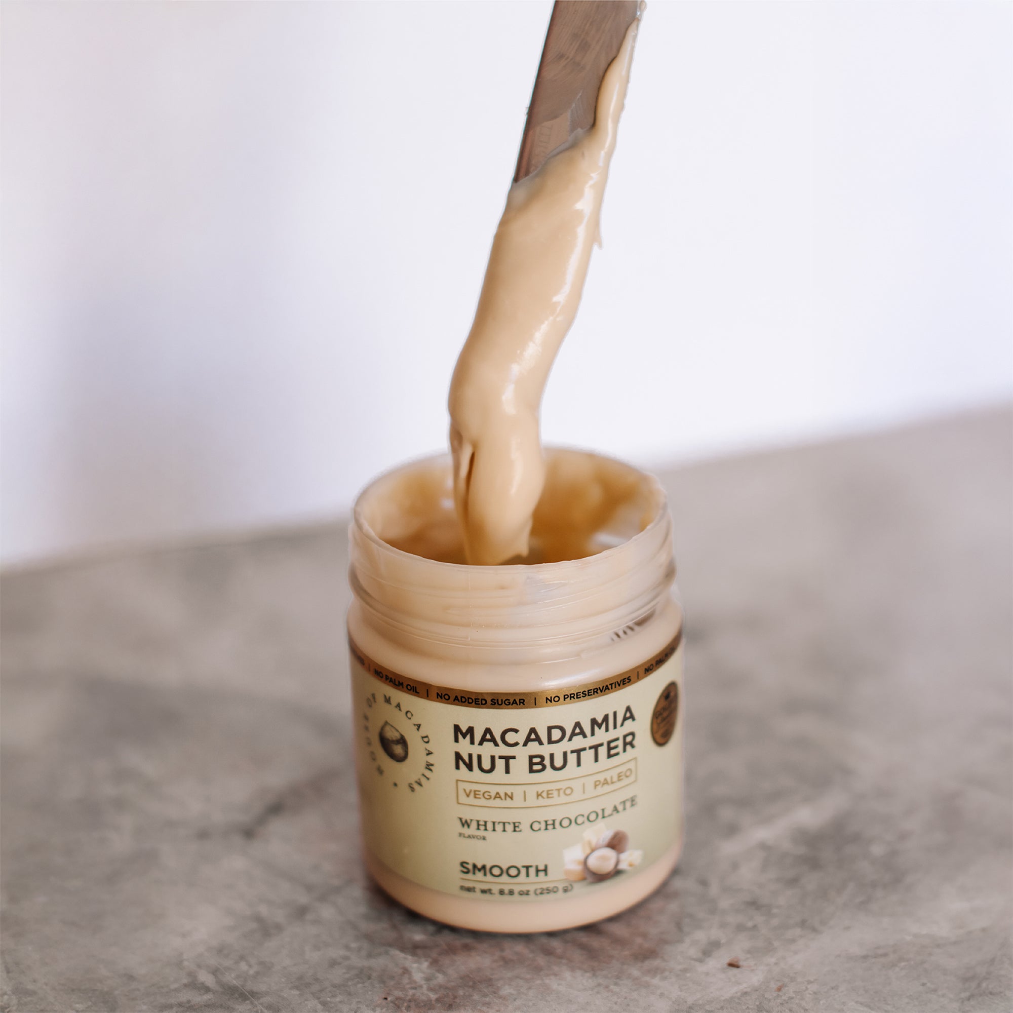 House of Macadamias White Chocolate Macadamia Nut Butter dripping from a knife held above the jar, showing creamy texture