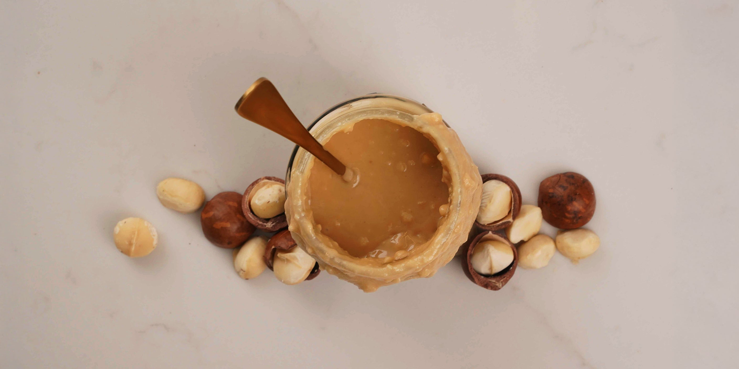 Jar of macadamia nut butter, the healthiest nut butter, on a marble table top surrounded by macadamia nuts in shells and shelled.