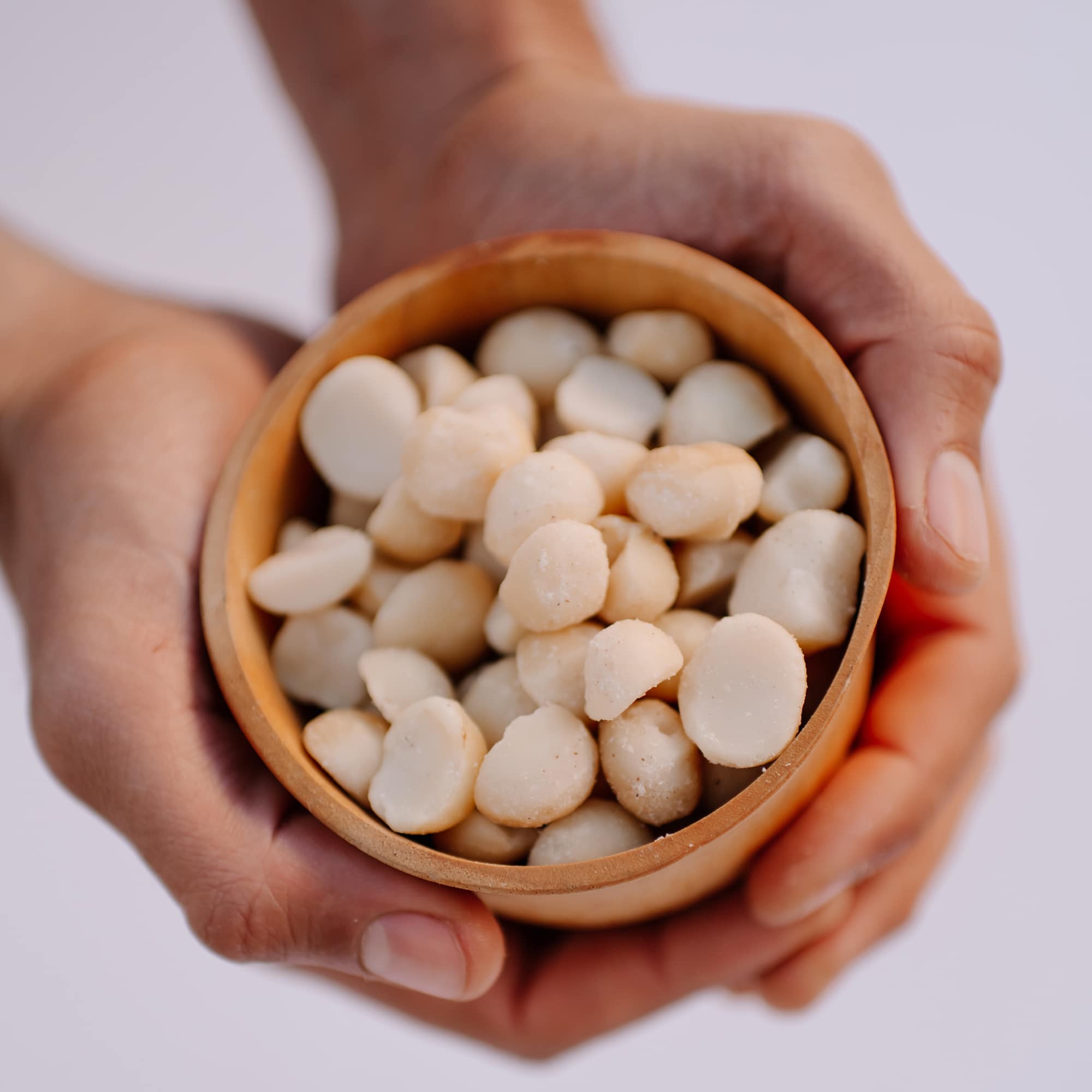 Raw macadamia nuts held in hands in a wooden bowl.