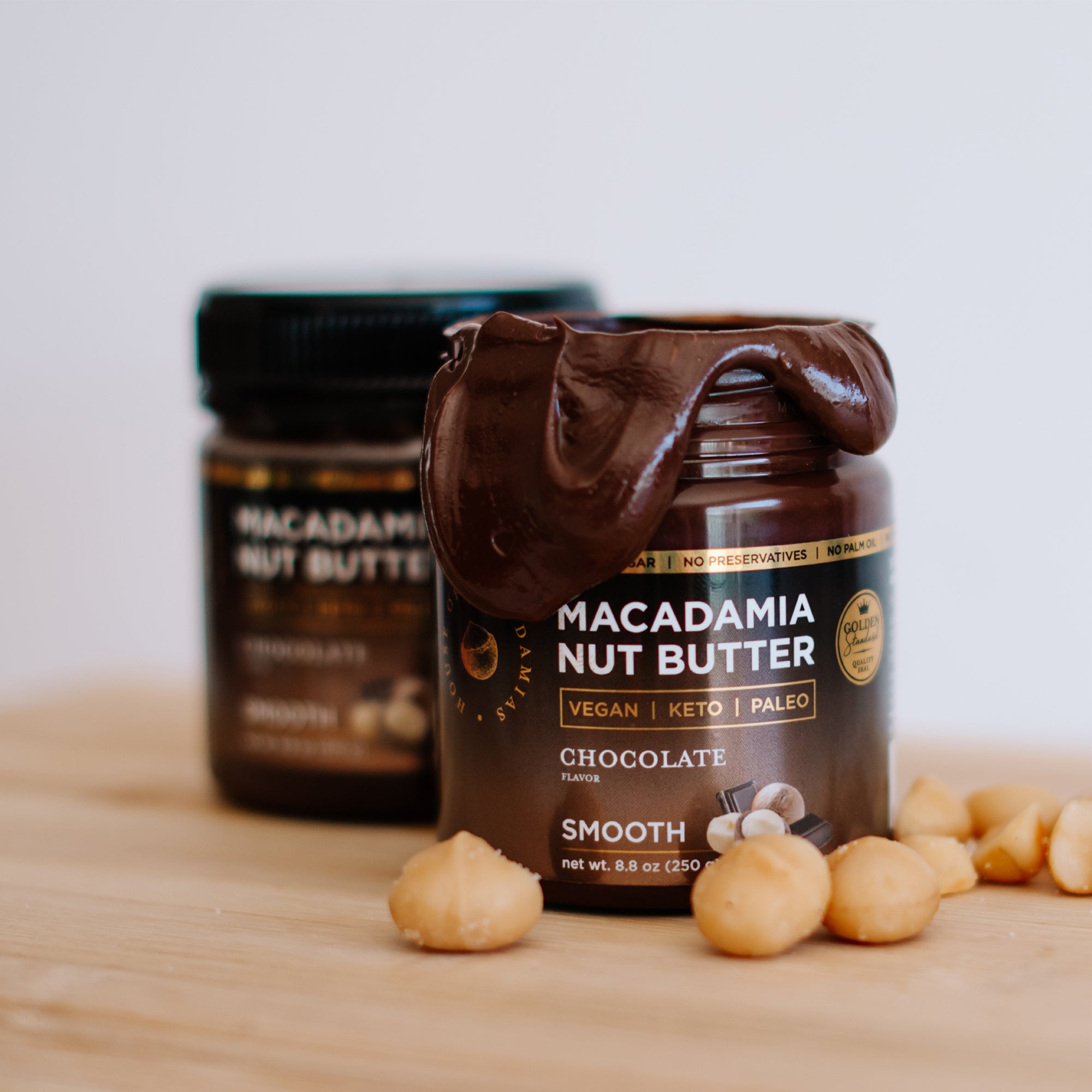 House of Macadamias Chocolate Macadamia Nut Butter jars, one open with creamy nut butter smeared around the lid, fresh macadamia nuts in the foreground — a natural, healthy spread rich in monounsaturated fats