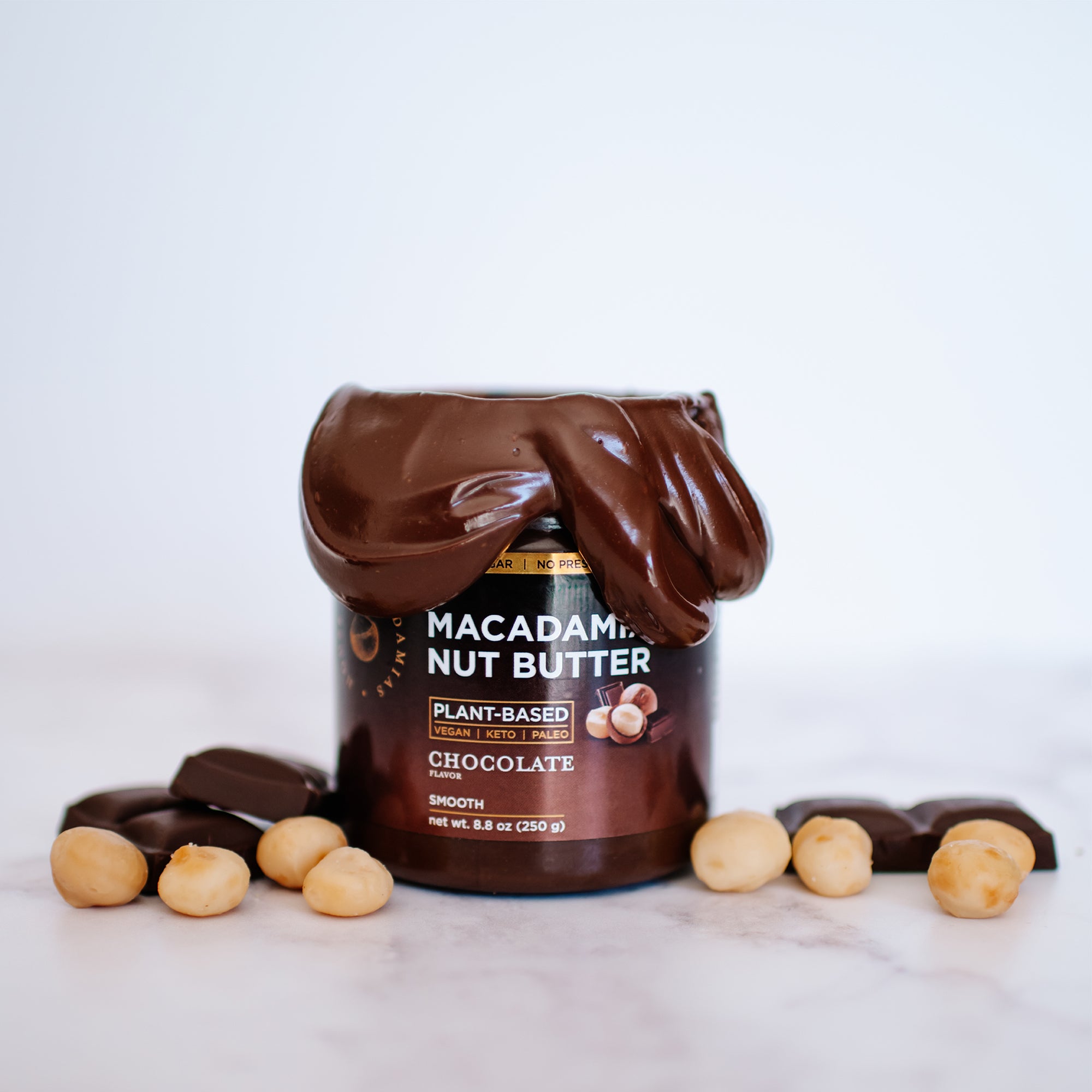 House of Macadamias Chocolate Macadamia Nut Butter jar front view with lid open, creamy nut butter around the sides, and macadamia pieces and chocolate chunks beside the jar — a natural, indulgent, and healthy spread