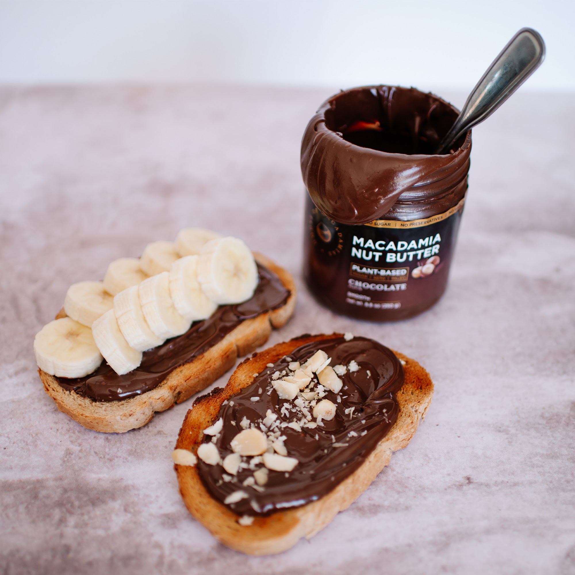House of Macadamias Chocolate Macadamia Nut Butter jar next to two slices of sourdough toast — one topped with crushed macadamia nuts and the other with banana slices, making a nutritious and wholesome breakfast or snack