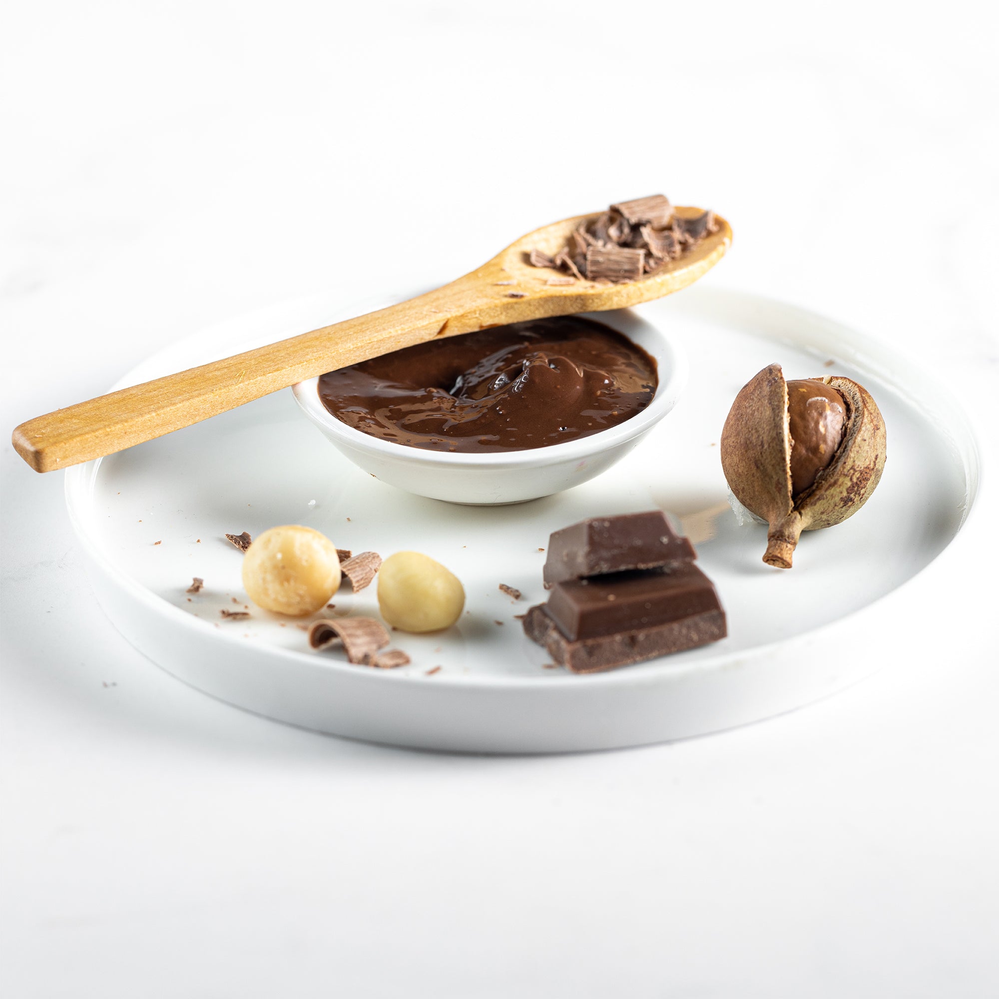 House of Macadamias Chocolate Macadamia Nut Butter served in a bowl on a plate, with chocolate pieces on a spoon above it, surrounded by whole macadamia nuts and an un cracked macadamia shell — a natural and indulgent treat