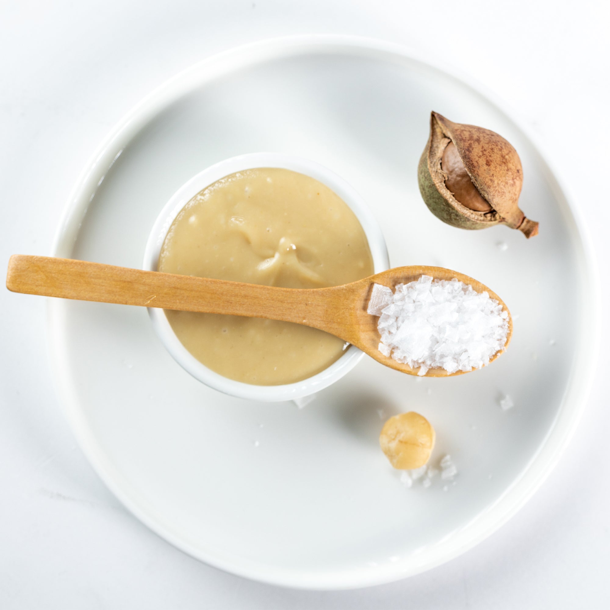 All Natural Macadamia Nut Butter in a bowl with sea salt, a whole macadamia nut, and an un cracked macadamia shell on the same plate