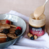 House of Macadamias Sea Salt Macadamia Nut Butter jar with macadamia nuts beside it, and nut butter drizzled over pancakes topped with blueberries and raspberries