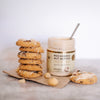 House of Macadamias White Chocolate Macadamia Nut Butter jar next to macadamia nut cookies drizzled with white chocolate macadamia butter, with macadamia nuts in the foreground — a delicious, natural treat